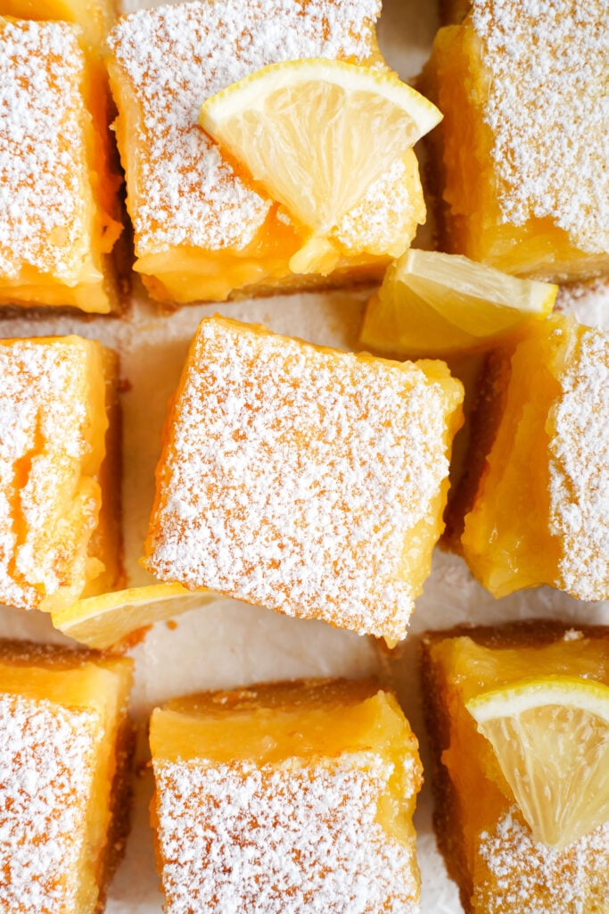 Gros plan aérien de barres au citron surmontées de sucre glace et garnies de quartiers de citron sur du papier sulfurisé.