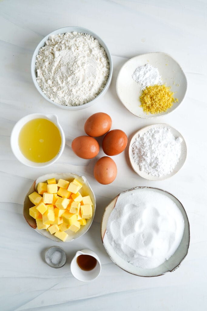 Ingrédients pour la recette de barres au citron affichés sur une surface blanche, y compris de la farine, du sucre en poudre, du beurre, des œufs, du jus de citron, du zeste de citron, du sucre glace, du sel, de l'extrait de vanille et de la levure chimique.