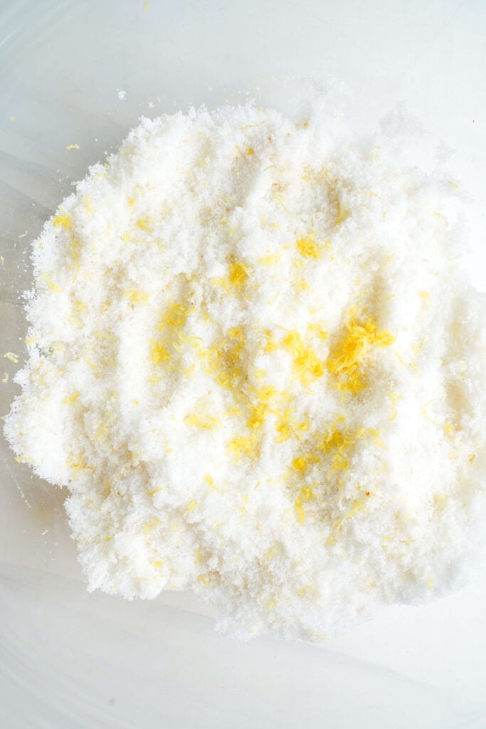 Sucre granuleux mélangé avec du zeste de citron frais dans un bol, montrant l'étape où les huiles d'agrumes sont libérées pour améliorer la saveur des barres au citron.