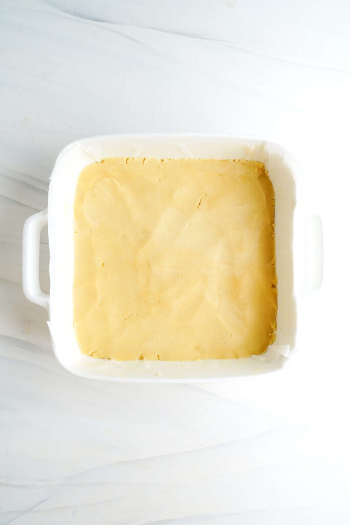 Croûte de sablé uniformément pressée dans un plat de cuisson de 9x9 tapissé de papier sulfurisé, prête à être cuite pour les barres au citron.