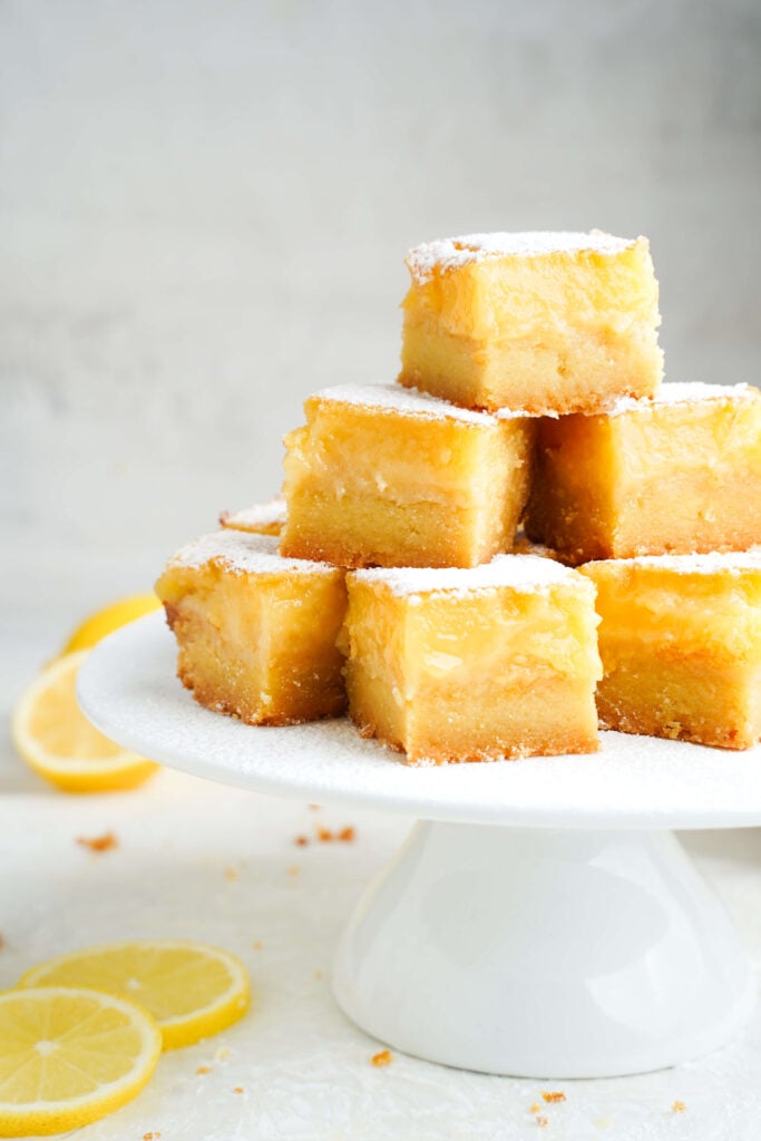 Barres au citron empilées sur un support à gâteaux blanc, saupoudrées de sucre glace et entourées de tranches de citron frais.