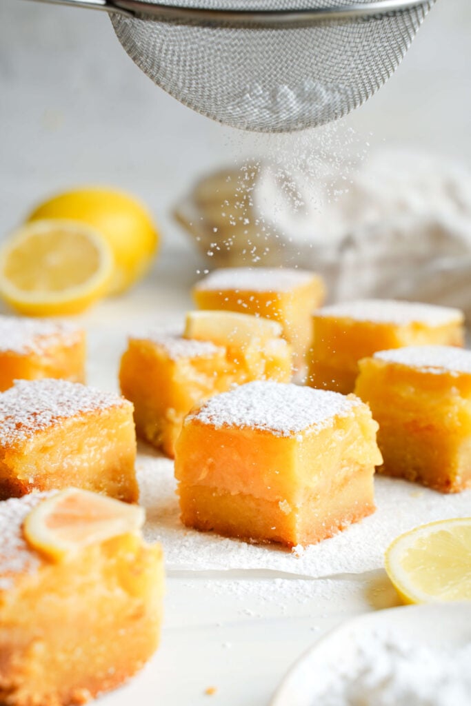 Gros plan de sucre glace saupoudré sur des barres au citron avec un tamis en maille, mettant en avant la texture douce et dorée.