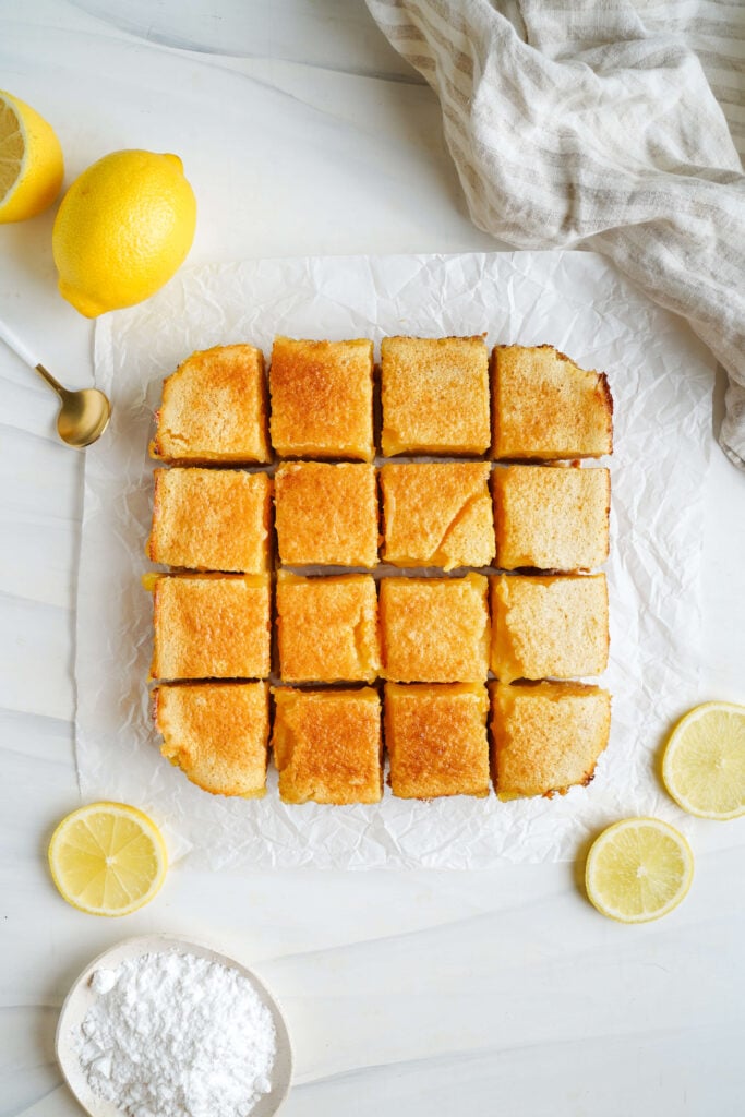 Barres au citron fraîchement cuites et réfrigérées, coupées en carrés sur du papier sulfurisé, entourées de tranches de citron et de sucre glace.