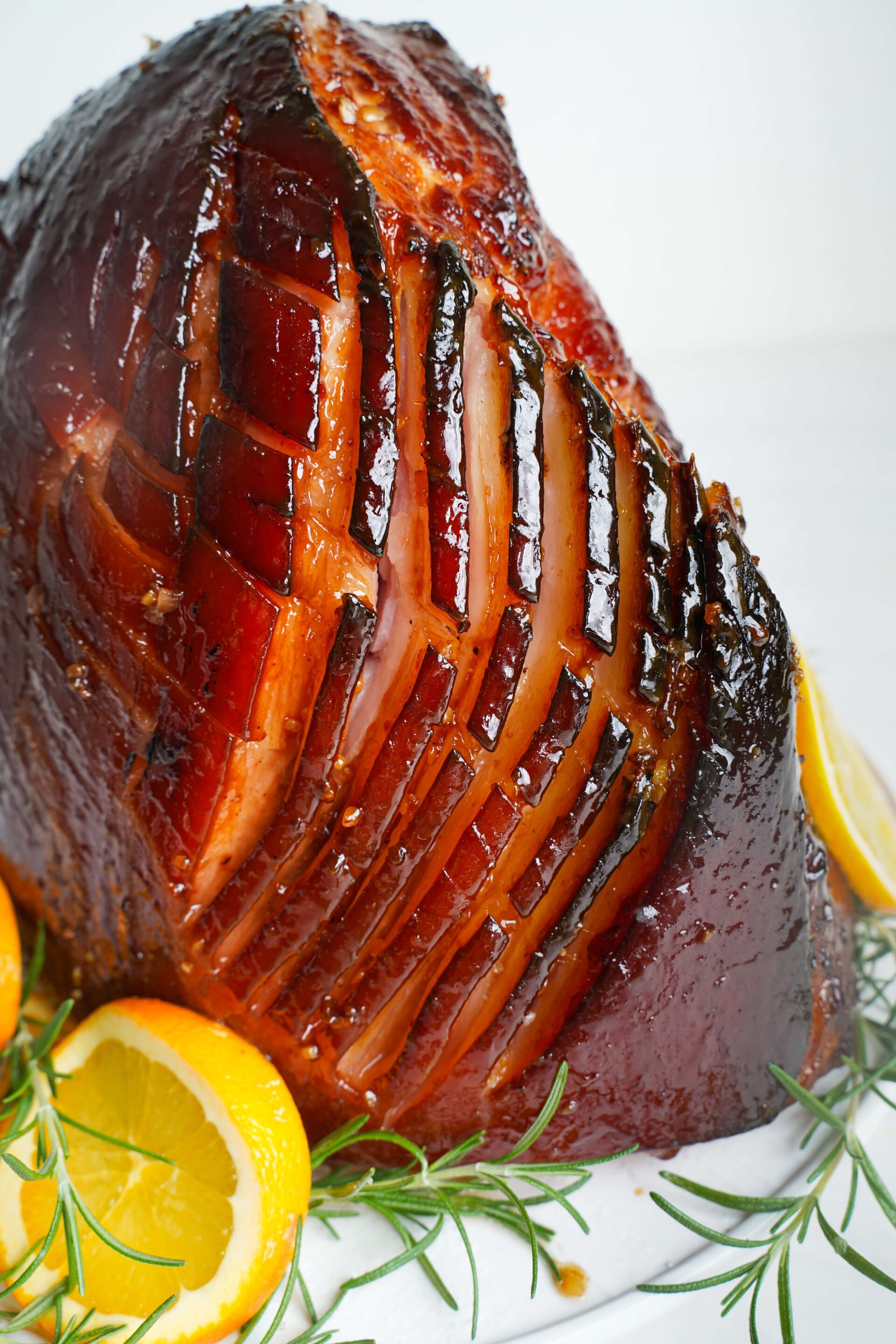 easy-brown-sugar-glazed-ham Jambon glacé au sucre brun entier tranché et servi avec des tranches d'orange et du romarin sur un plat blanc.